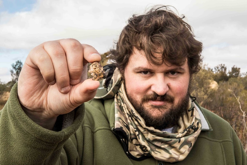 A man with a beard holding a small gold nugget in his fingertips