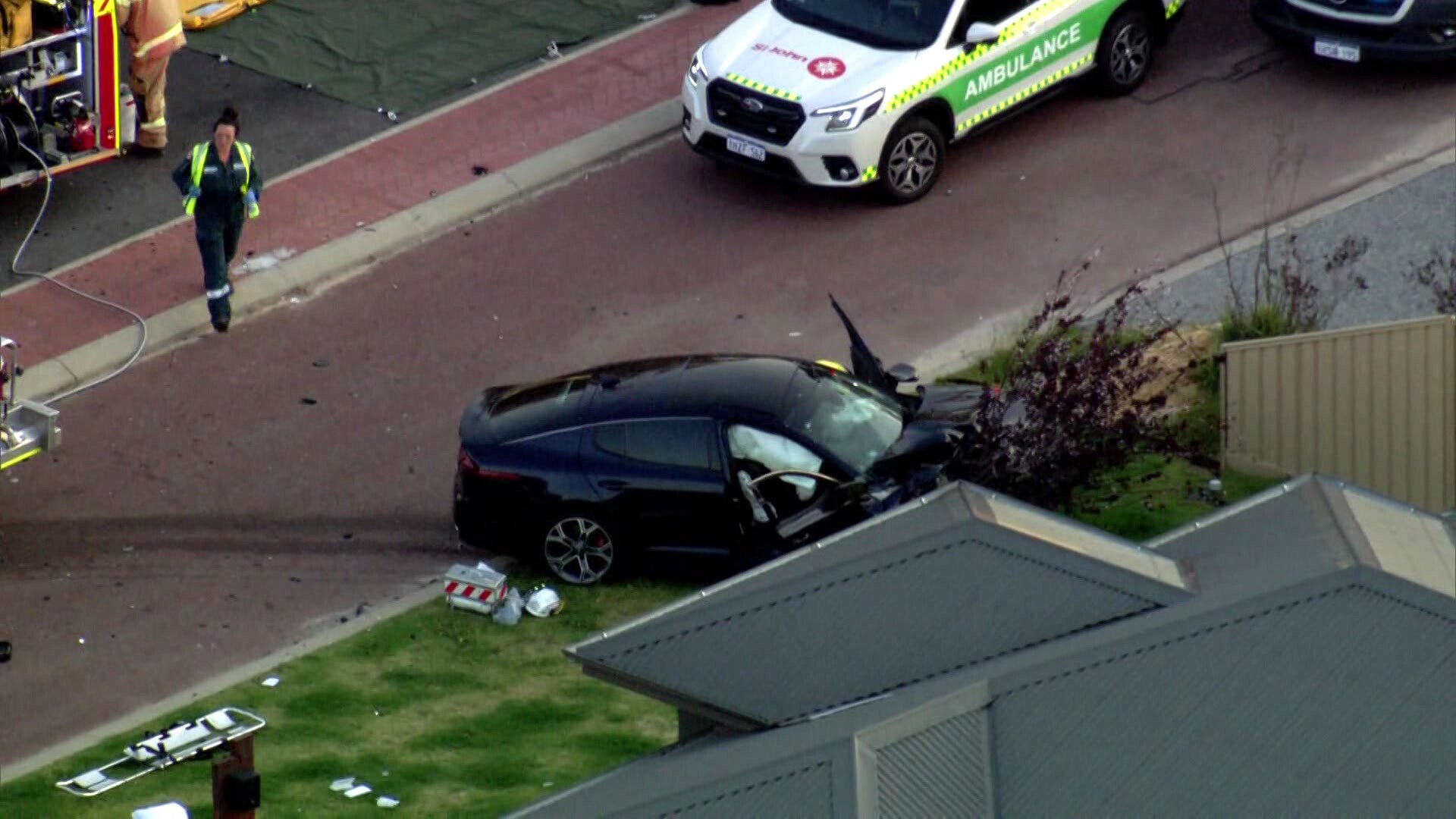 The wreckage of a black sedan parked on a front lawn of a house after veering off the road in a crash, with an ambulance near.