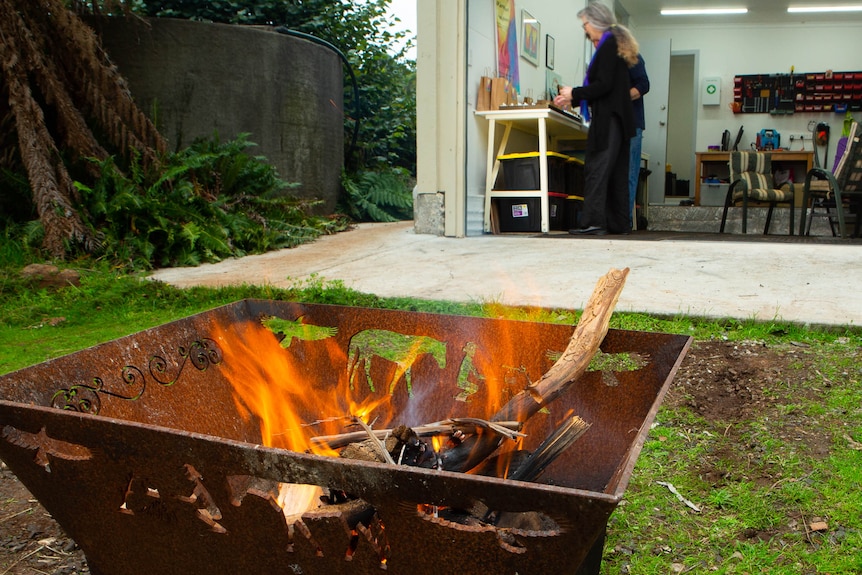 A firepot with orange flames in foreground, people working in shed behind.