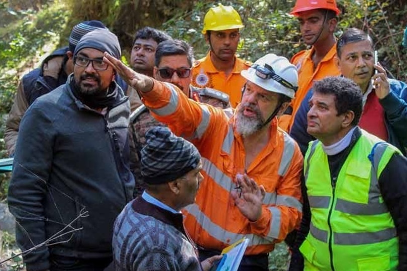 11 men stand in a tree-filled forrest. One man who has a beard is in the centre wearing hi-vis gear and helmet and pointing 