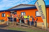A group of smiling people in fluorescent clothing stand on steps in front of orange building.