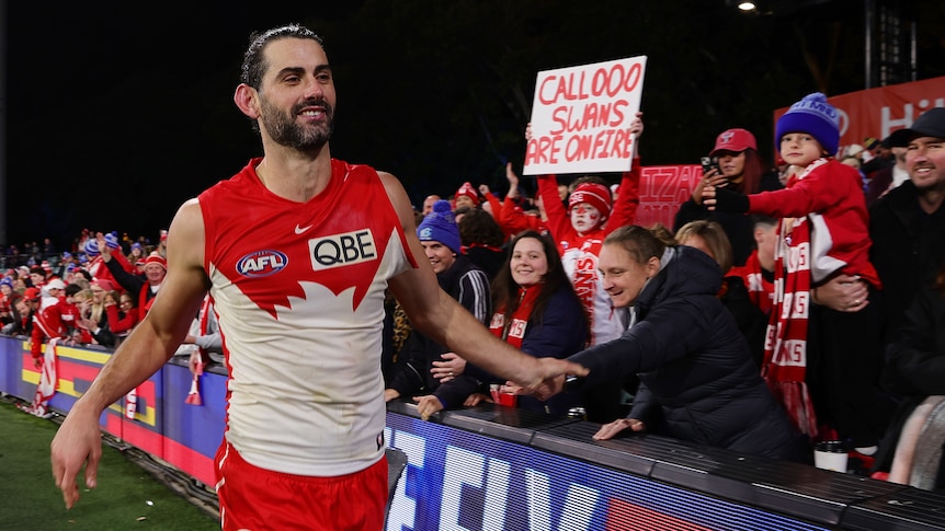 A smiling Sydney Swans player reaches out to fans after a win, with a sign in the background 'Call 000 Swans are on fire'. 