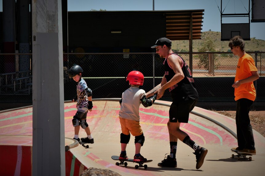 An adult guides a kid with a red helmet along at skateboard course