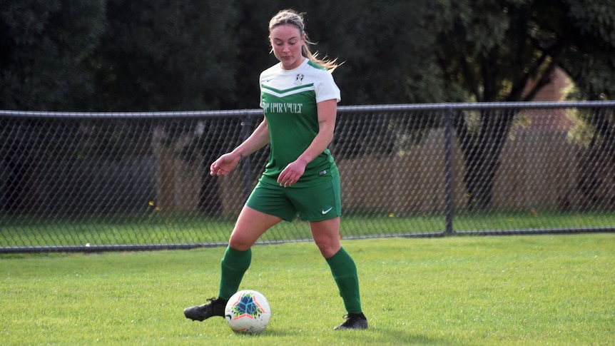 A blonde woman dribbling a soccer ball in the middle of a field