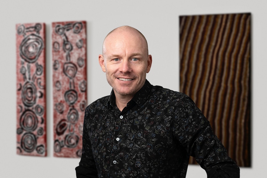 A bald man smiles in front of artwork.