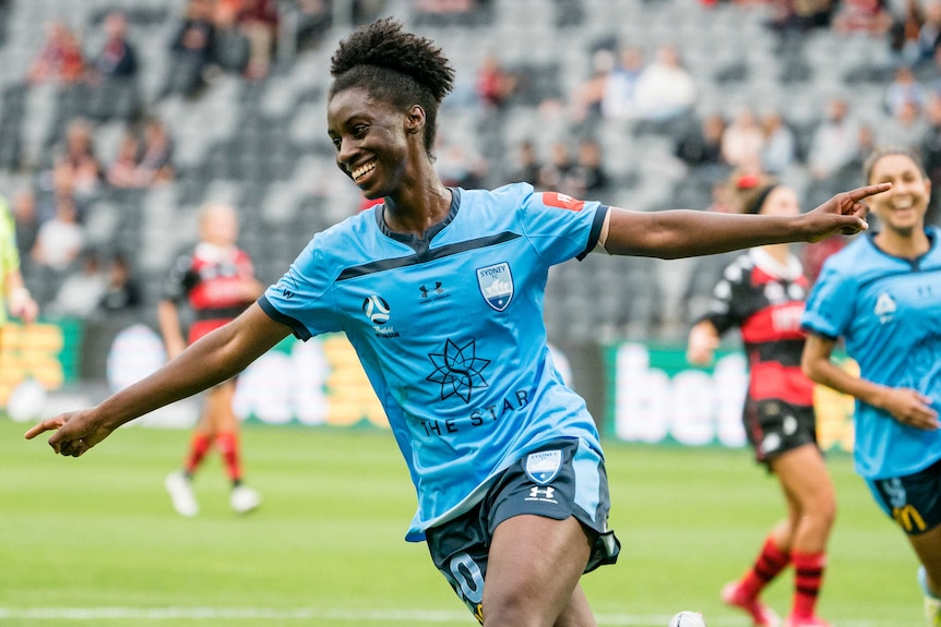 A female football player smiles and holds her arms out to celebrate a goal.