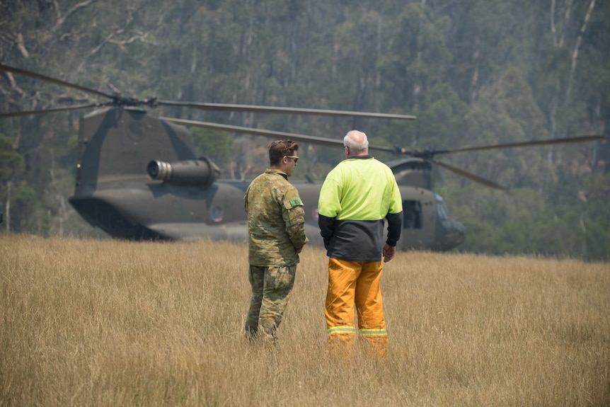 A soldier stands in a field with a middle-aged man with an army helicopter in the background