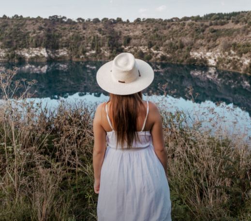 A woman wearing a white dress and hat looking over a blue-ish lake with reeds and cliffs.