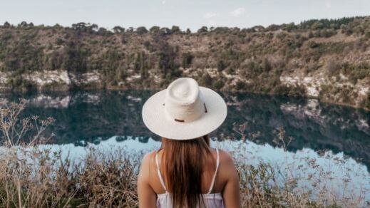 A woman wearing a white dress and hat looking over a blue-ish lake with reeds and cliffs.
