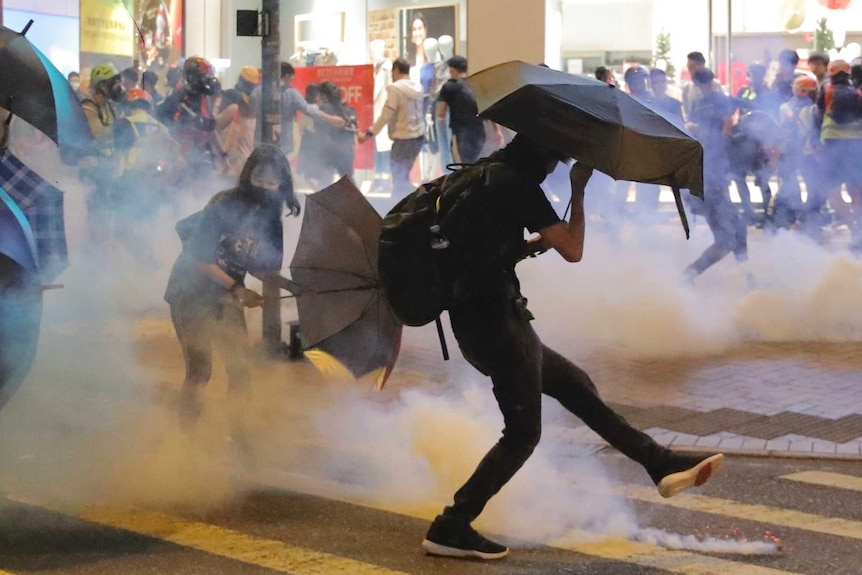 People carrying umbrellas and wearing face masks near a Marks and Spencer shop kick a smoking teargas shell away