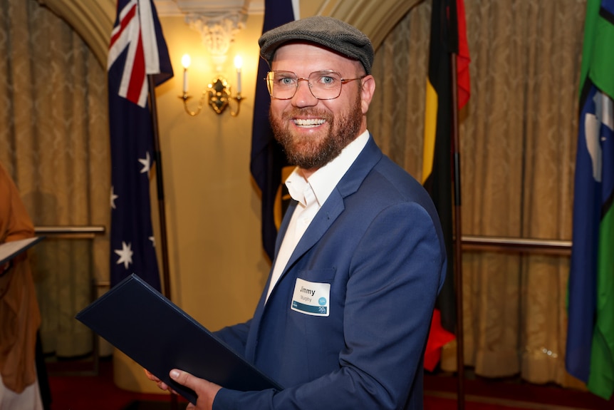 A man wearing a short-brimmed cap smiles inside a building with flags in the background.