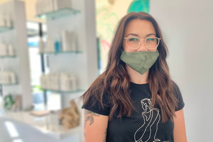 A woman with long brown hair, wearing a mask and glasses smiles in a hair salon
