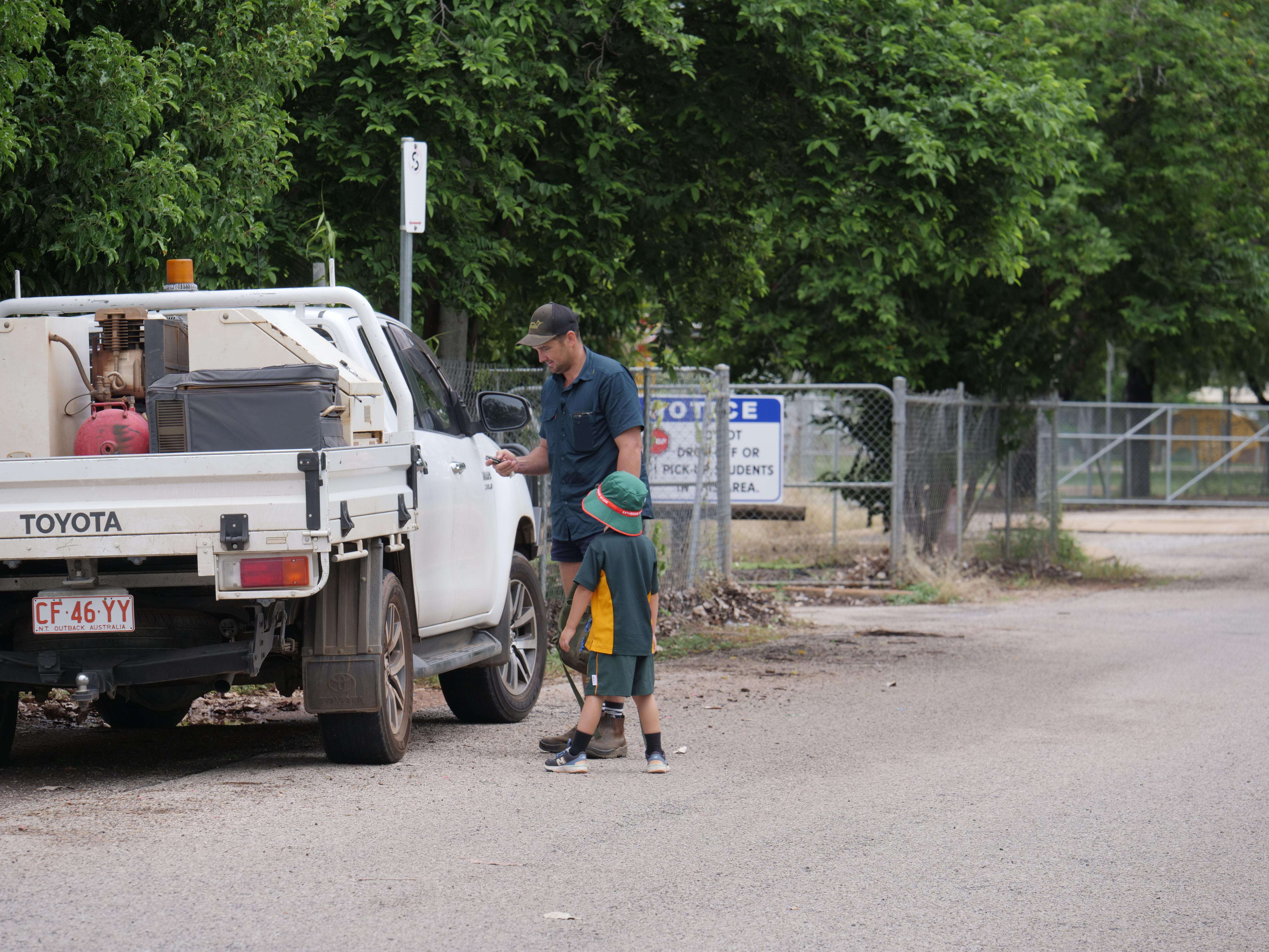 A young child in a green and yellow school uniform prepares to get in a grey ute with his dad at school pickup. 