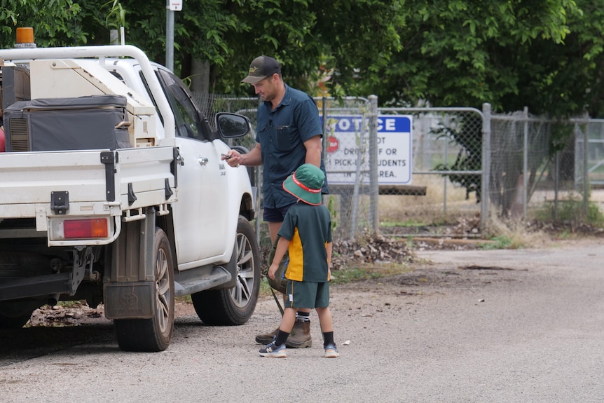 A young child in a green and yellow school uniform prepares to get in a grey ute with his dad at school pickup. 