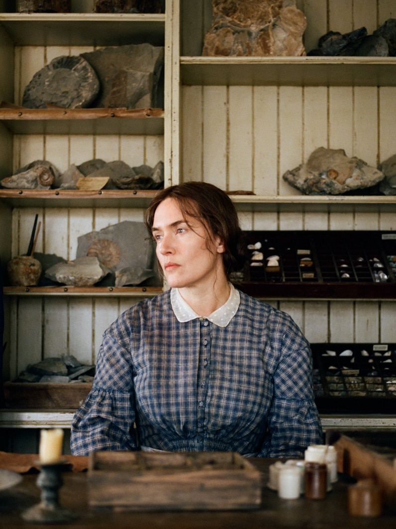 Kate Winslet wearing 19th century dress sits at desk with fossils on shelving behind her, turning left and staring into distance