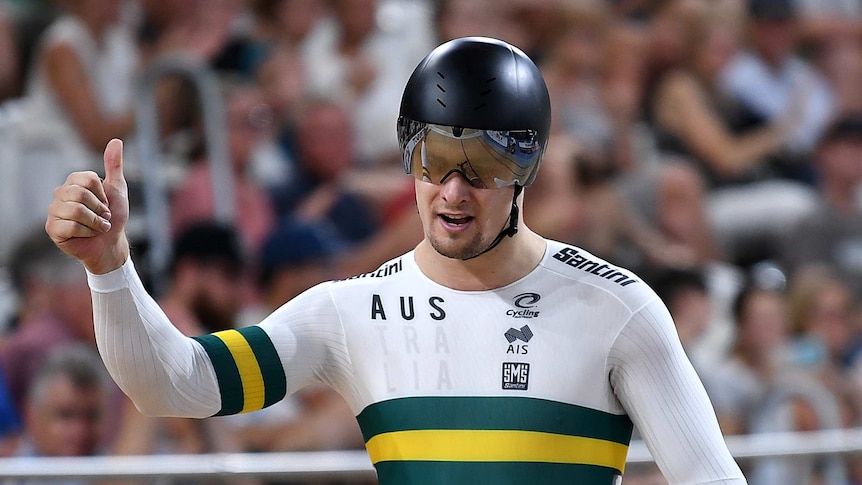 Matthew Glaetzer holds his thumb up, wearing a helmet and white track cycling suit with a gold and green band across the chest