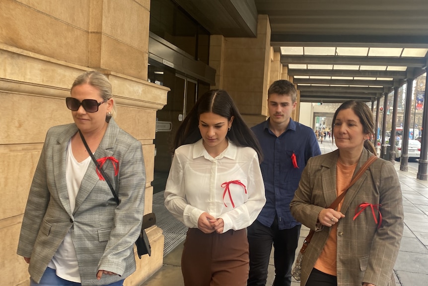 A young woman walks outside a courthouse with two elderly women and a young man.  They all wear red bows tied to their tops. 