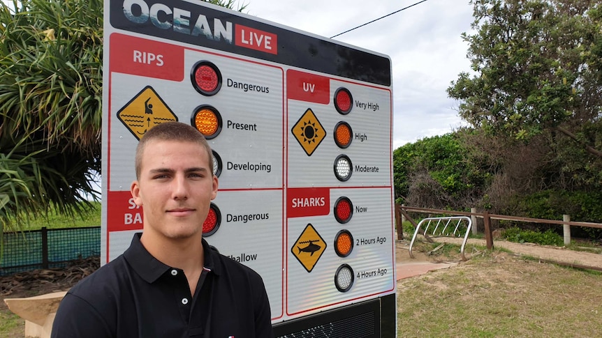 A young man stands in front of a beach signs warning about dangerous currents