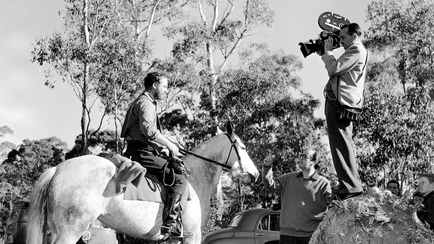 Black and white photo of man on a horse, and another man nearby holding a video camera facing the horse. Bush surrounds them.
