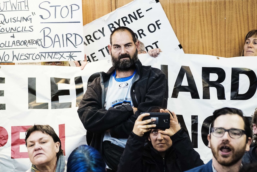 A angry-looking protester stands with his arms cross in front of placards at the council meeting.