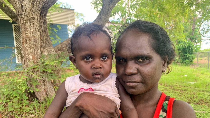 Bronwyn Bianamu holding her child, looks at the camera. She is standing outside, surrounded by trees.