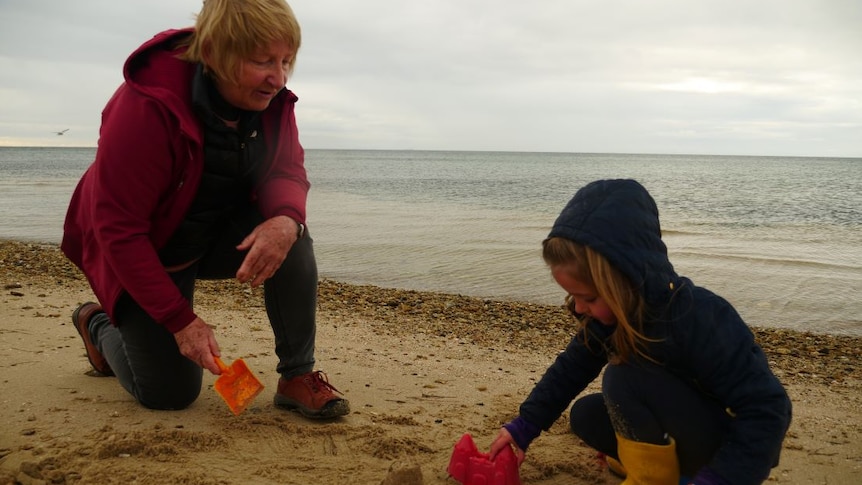 A grandmother with short blonde hair builds sandcastles with her 4yo granddaughter on a cold, overcast day.