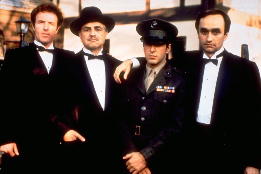 James Caan standing in a black suit as character Sonny in The Godfather, standing beside Marlon Brando, Al Pacino, John Cazale.