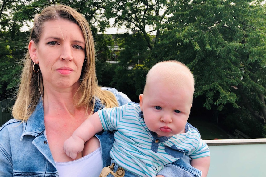 Jennifer Roe looks to the camera with a neutral expression on her face as she holds her baby on her right. They both wear blue.