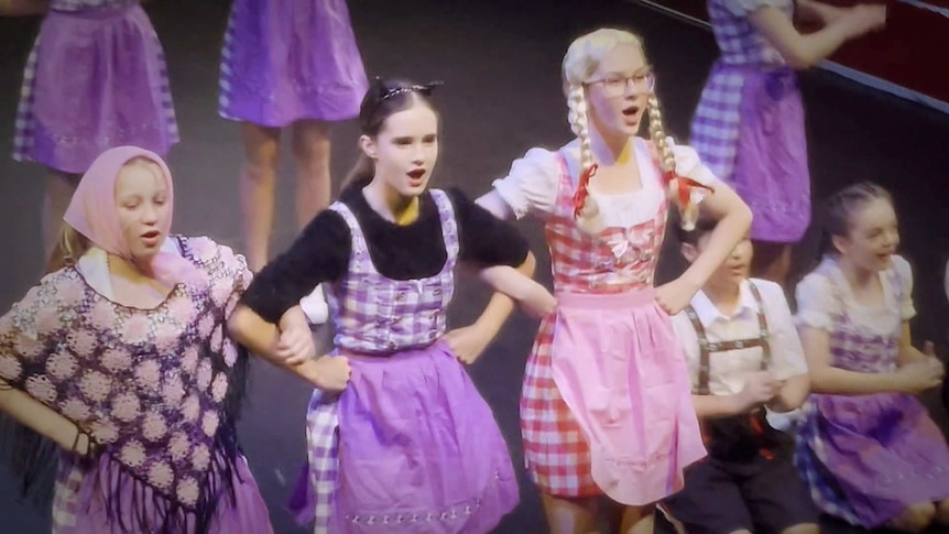 Three teenagers in sound of music style dresses and wigs linking arms and singing and dancing on stage.