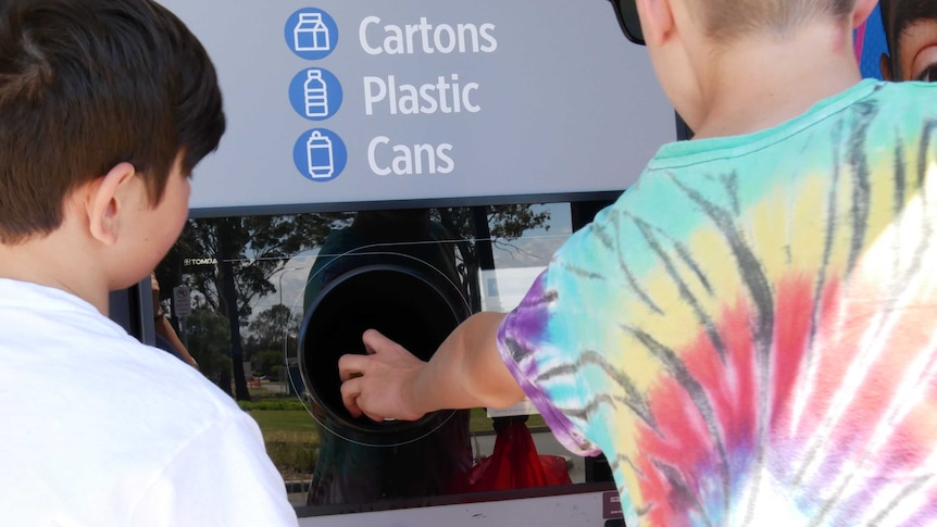 Two kids putting bottles into the return and earn chutes. Photo taken from behind so you can only see their backs.