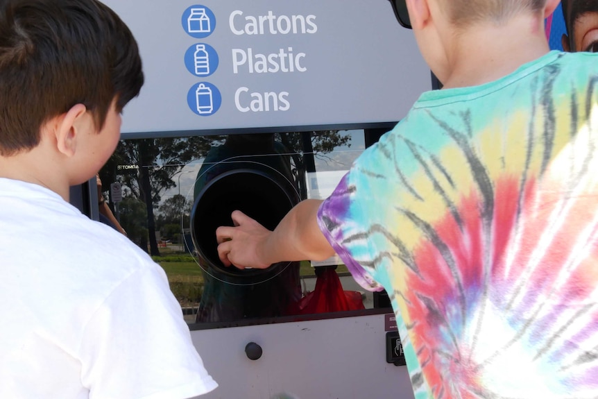 Two kids putting bottles into the return and earn chutes. Photo taken from behind so you can only see their backs.