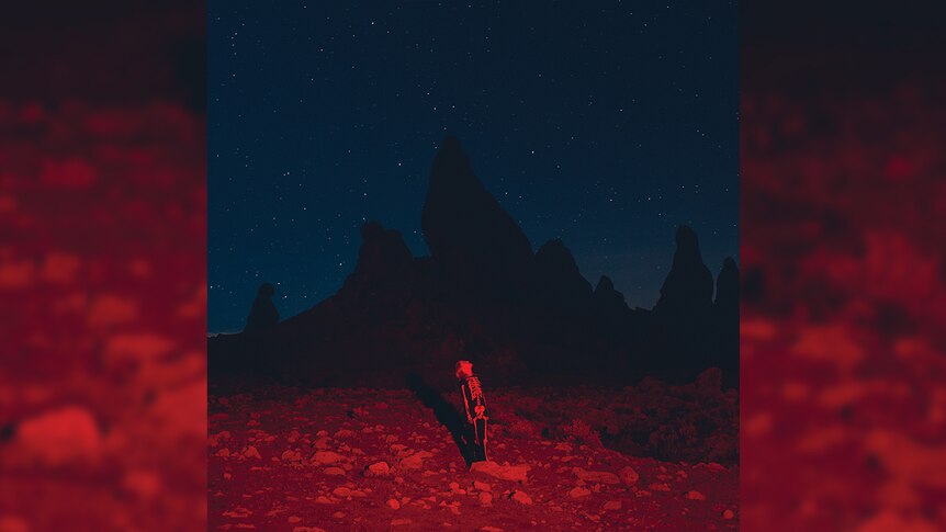 Phoebe Bridgers stands in the middle of the desert at night, wearing a skeleton suit. A red light is cast upon her.