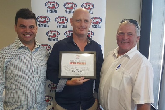 A man stands in front of an AFL banner holding an award