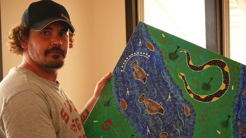 Aboriginal Artist Dean Horsburgh holds an artwork depicting a river scene with snake, fish and emus.