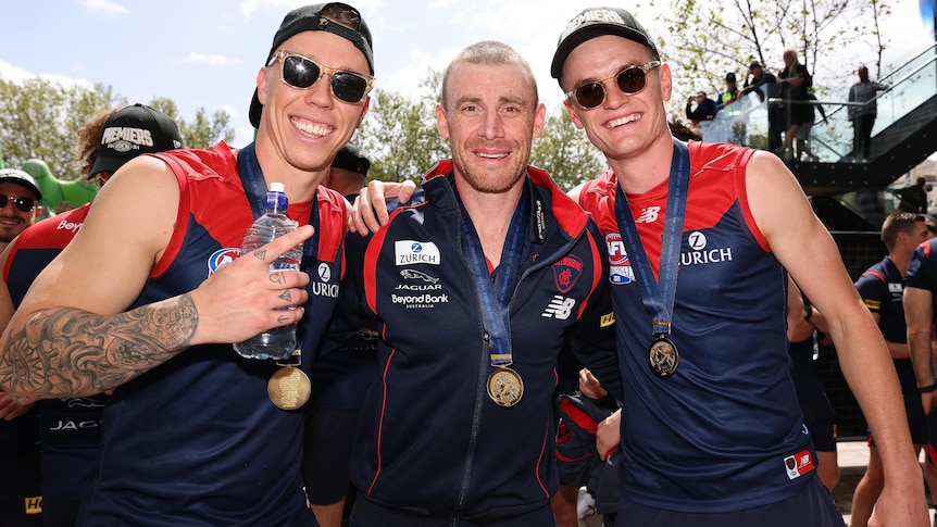James Harmes, Simon Goodwin and Bayley Fritsch stand arm in arm. All have medals, and the two players are wearing sunglasses