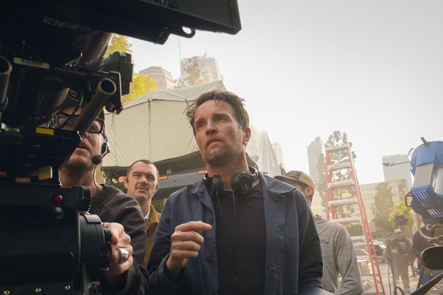 A white man with brown hair and stubble behind the scenes on a movie set, looking at a camera shot