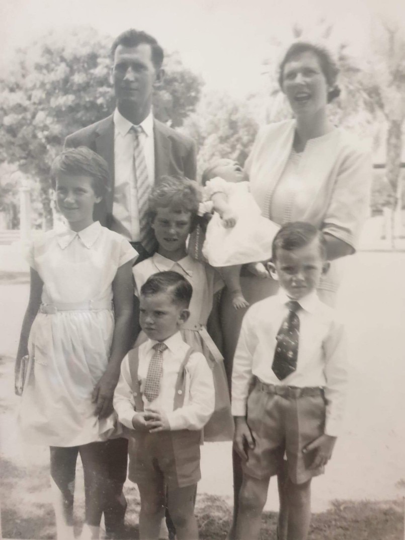 A black and white photo of a couple with four young children, the woman is holding a baby, standing outdoors.