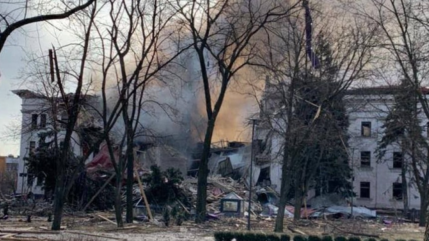 A large white building left in ruins and billowing smoke.