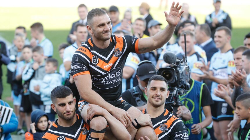 A male NRL player is chaired from the field on the shoulders of two teammates as he waves to the crowd with his left hand.