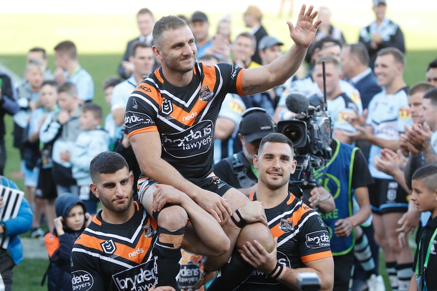 A male NRL player is chaired from the field on the shoulders of two teammates as he waves to the crowd with his left hand.