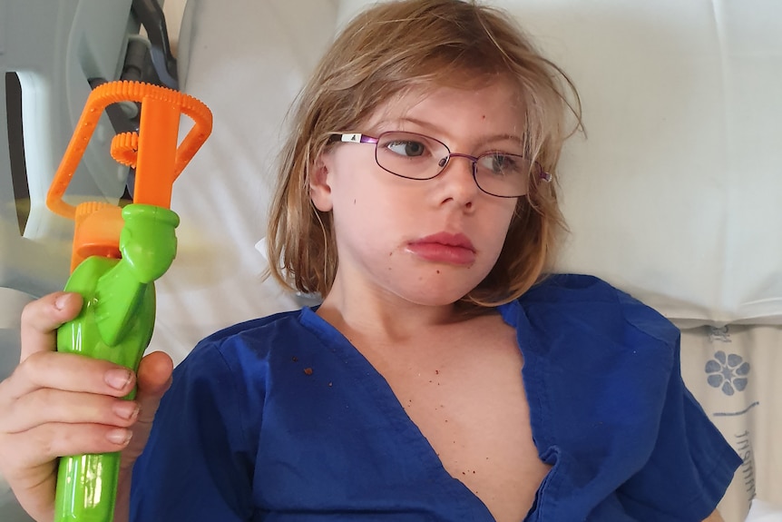 A forlorn looking young girl in hospital bed, wearing blue hospital gown, holds a green and orange equipment, wears glasses.
