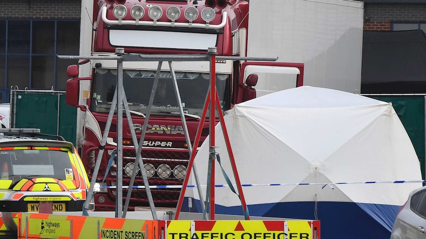 A truck with a red cab and a white body is seen behind a metal structure. A police vehicle with a trailer is seen in front of it