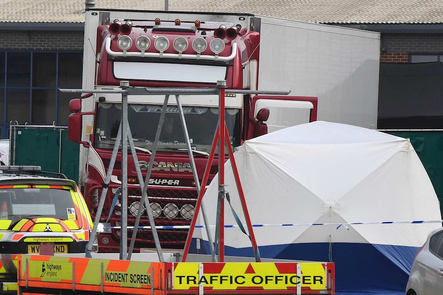 A truck with a red cab and a white body is seen behind a metal structure. A police vehicle with a trailer is seen in front of it