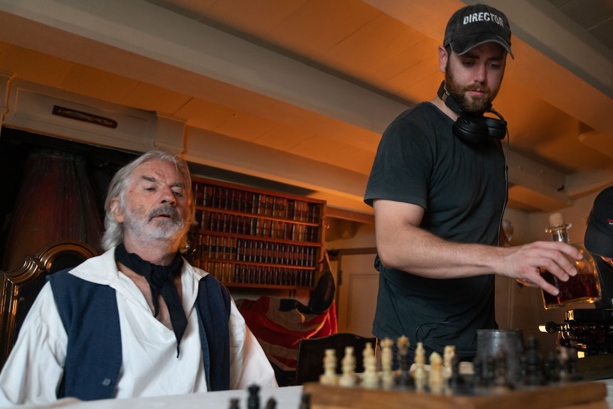 Nathan Colquhourn prepares a scene inside with a chess board on a table and John Jarratt sitting at the table.