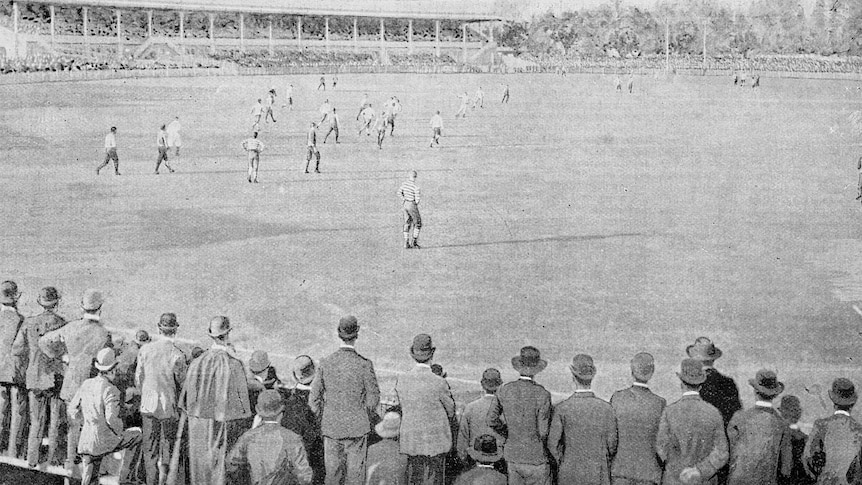 Black and white photograph behind crowd watching football players.