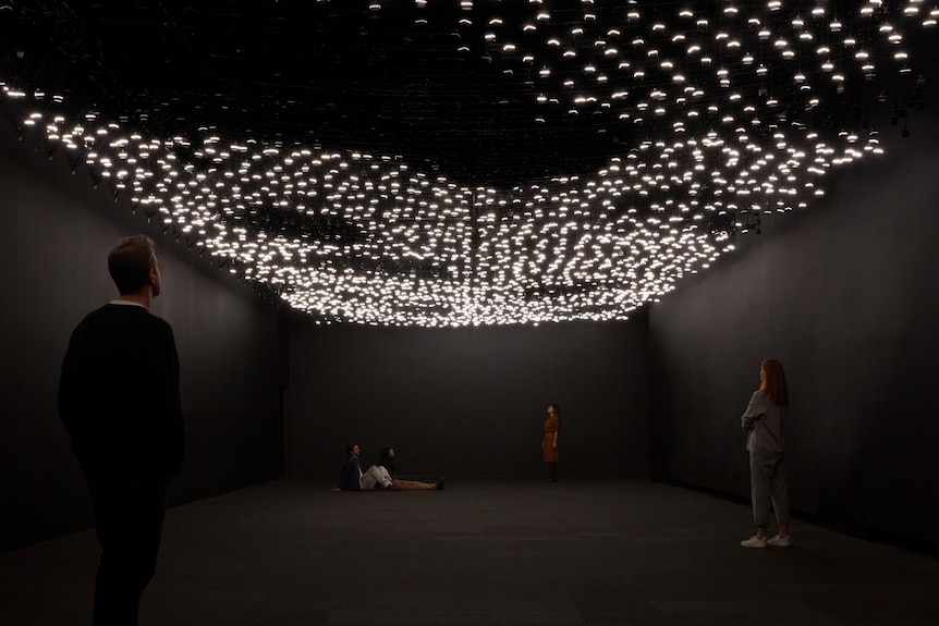 Four people stand in a dark room, under a canopy of thousands of speakers with little lights attached.