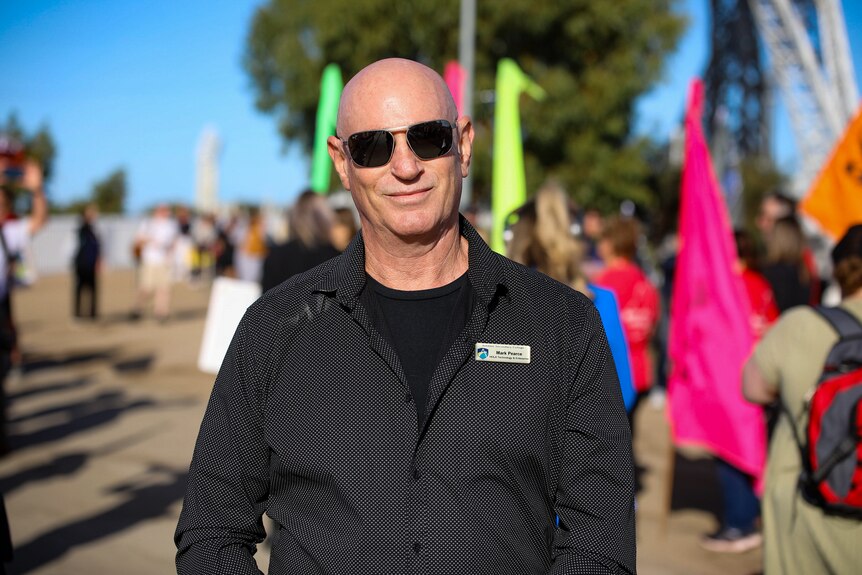 A mid-shot of teacher Mark Pearce outdoors at a rally wearing a black shirt and sunglasses, posing for a photo.