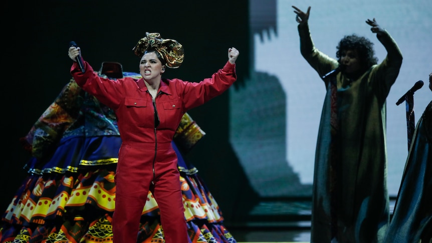 A woman stands on a stage pulling a fierce face with both hands in the air. She wears a red boilersuit and her hair is wrapped.