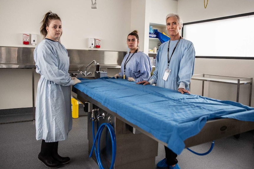 Morgue workers stand around a metal dissection table