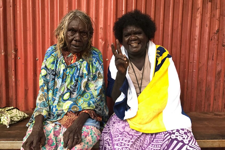 Two women from the Tiwi Islands sitting in front of red corrugated iron wall.